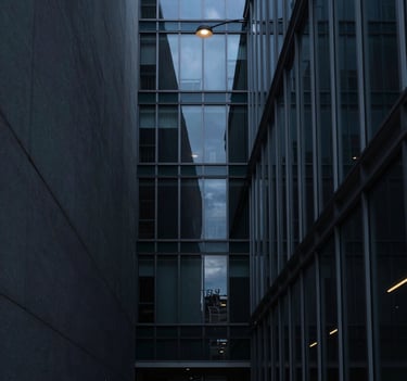 A minimalist vertical photography shot of a narrow alleyway between modern glass buildings. The palette features deep slate and muted blue tones. A single overhead lamp provides a pool of soft light against the dark shadows. International / Western metropolitan setting.