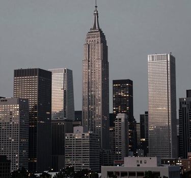 Atmospheric photography of a North American / US urban skyline at dusk. The buildings are rendered in silver grey and charcoal black, with the first soft off-white lights of the evening beginning to glow against a cool grey sky.