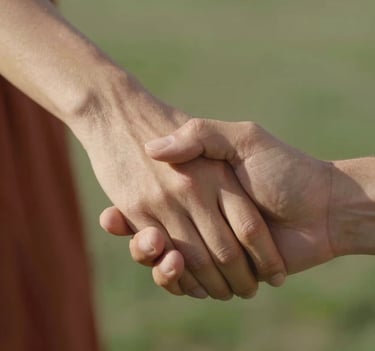 A close-up photograph of a couple's hands gently intertwined, focusing on the texture of their skin and the soft glow of a North American afternoon sun. The background is a soft blur of terracotta and sage green colors, emphasizing the intimacy of the touch.