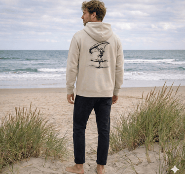 A man in a beige graphic hoodie with a wing foil design stands on a sandy beach looking at the ocean.