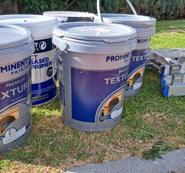 Prominent Paints exterior textured paint buckets and a step ladder on a lawn for a home painting project.