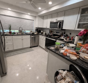 Messy modern kitchen with white cabinets, stainless appliances, and a full trash can.