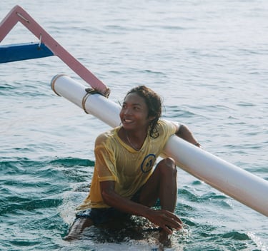 A smiling young Indonesian surfer resting on his board next to a traditional outrigger boat in the ocean.