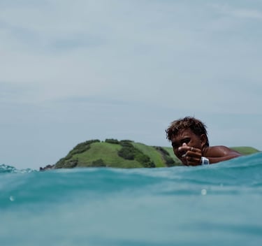 A smiling surfer gives in blue ocean water with a tropical green hill background.