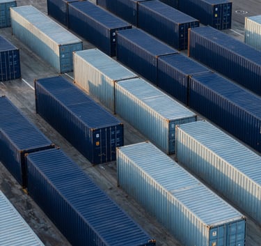 Aerial view of a clean, modern logistics hub in a Lusophone Internacional coastal city. Neatly arranged shipping containers in shades of Dark Navy Blue and Soft Steel Blue. Morning sun, high-contrast, minimalist architectural composition.
