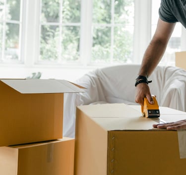 A man using a tape dispenser to seal cardboard moving boxes in a bright living room.