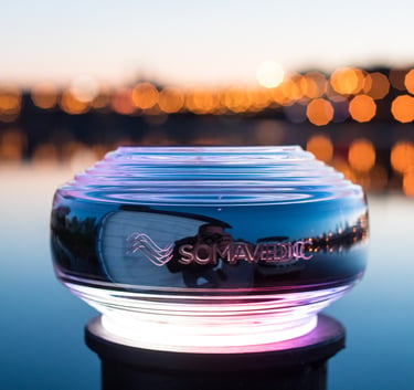 Glowing Somavedic EMF mitigation device outdoors at sunset with bokeh city lights background.