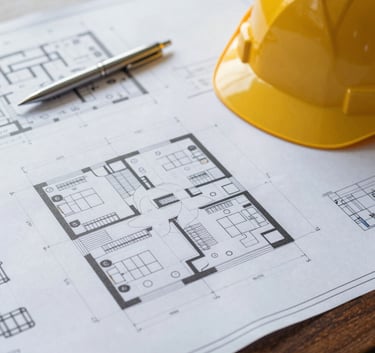 Close-up of high-quality architectural blueprints laid out on a dark mahogany desk. A modern metal pen and a professional hard hat sit beside the plans. Sophisticated, warm indoor lighting highlighting the precision of the drawings. Subtly includes brand colors #A68A6B and #2C3E50.