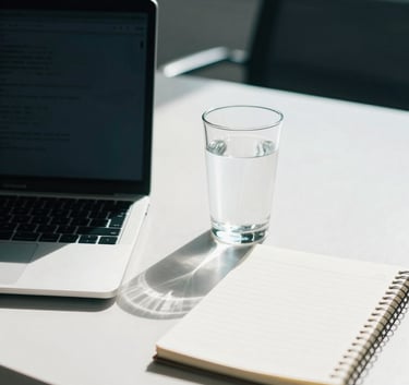 A professional desk setup in a sunlit Bothell, Washington office with a laptop, a clear glass of water, and a simple notebook. North American / US context with soft natural light and colors of white azure and dark teal.