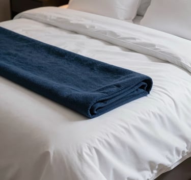Photography of a high-end apartment bedroom with perfectly made beds. The linens are crisp, soft white, and expertly tucked in. A midnight blue throw blanket is neatly folded at the end of the bed. The atmosphere is serene and inviting, suggesting a premium guest experience.