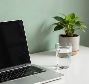 A clean, modern workspace with a sleek laptop and a glass of water. A single green plant sits in a ceramic pot. The lighting is bright and airy, with soft shadows and a wall painted in a gentle Sage Leaf green.