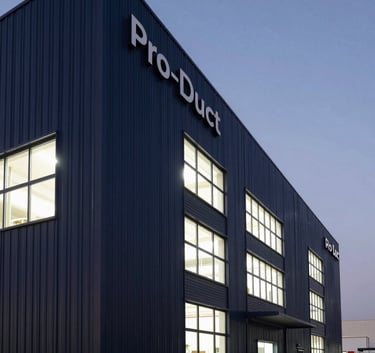 A low-angle architectural photograph of the Pro-Duct manufacturing plant in Riyadh. The modern facade features dark charcoal blue steel panels and bright industrial windows against a clear twilight sky.