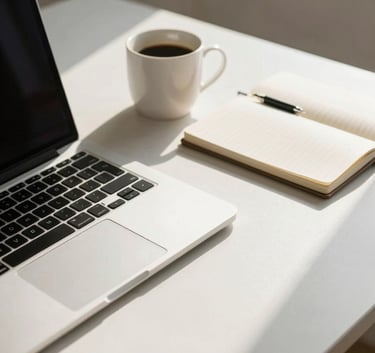 A minimalist workspace featuring a sleek laptop, a notebook, and a coffee cup on a soft white desk, illuminated by natural sunlight.