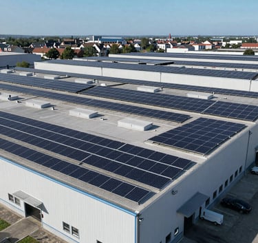 Wide aerial shot of a large industrial warehouse in a European / French commercial zone with a vast array of high-quality solar photovoltaic panels on the roof under a clear blue sky.