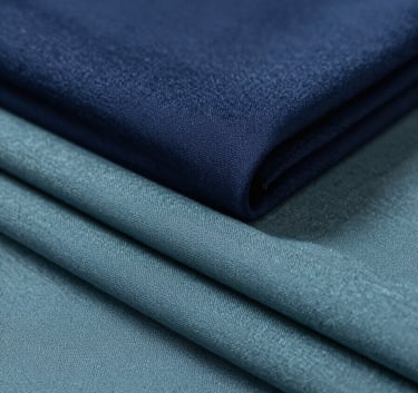 A professional and sophisticated studio shot of folded luxury fabrics in shades of navy blue #283645 and muted teal #5B7B7C. The composition is clean and minimalist, with soft lighting highlighting the fine texture of the weave.