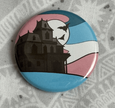 An image of a pin badge with a haunted house illustration in the colours of the trans flag.