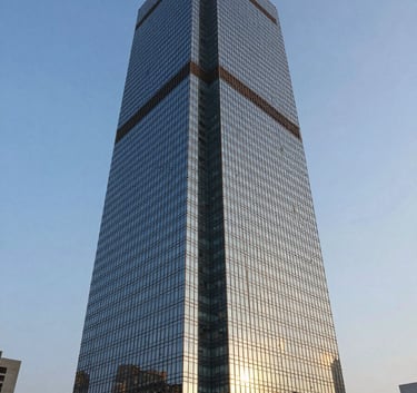 A sharp, wide-angle architectural photograph of a modern glass-fronted luxury skyscraper in Gurgaon under a clear slate blue sky. The building reflects the warm glow of sunset, conveying a sense of premium value and professional expertise.
