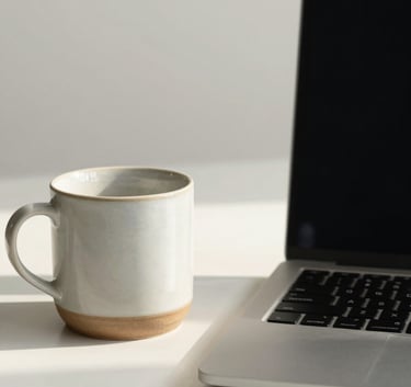 A minimalist North American / European workspace featuring a single ceramic cup and a sleek laptop on a soft off-white surface, bathed in natural morning light, creating a calm and professional atmosphere.