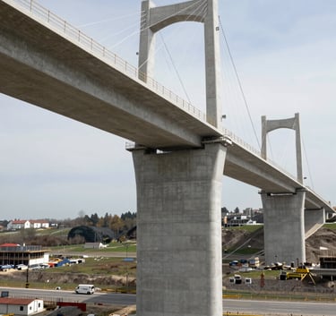 A professional photograph of a major civil engineering project, such as a bridge or highway under construction in a French / European landscape. The scene highlights precision engineering and solid silver grey concrete structures.