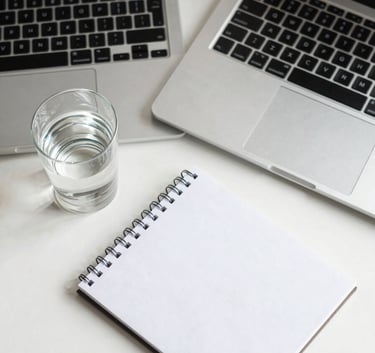 A top-down shot of a minimalist workspace with a thin silver laptop, a single glass of water, and a white notepad on a Pearl White surface, reflecting a clean and professional mood.