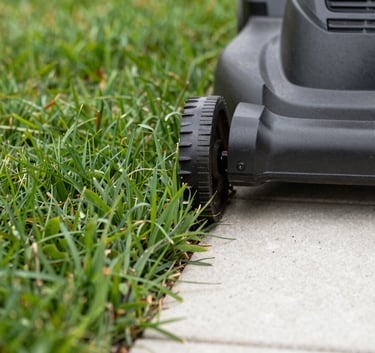 A close-up, low-angle shot of a professional lawn mower blade cleanly cutting through vibrant leafy green grass, with a sharp edge being created against a bone white stone walkway.