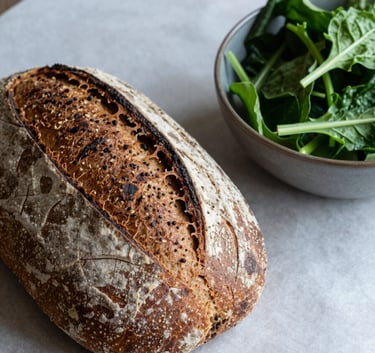 A cozy, Scandinavian-style food photograph featuring a rustic loaf of bread and a bowl of fresh greens on a crisp parchment surface. The lighting is soft, creating an approachable and high-quality artisanal feel. North American setting.