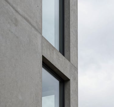 Close-up of a modern architectural detail, showing the intersection of a concrete wall and a glass pane against a cloud white sky, emphasizing precision and clean lines.