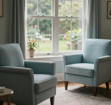 A peaceful and inviting therapy room in a US setting, featuring two comfortable upholstered chairs in a light sky blue, a soft rug, and a large window overlooking a calm garden. The atmosphere is quiet and reassuring.