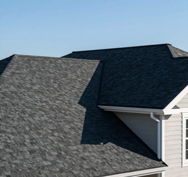 A wide-angle professional photograph of a beautiful North American suburban house featuring a newly installed dark asphalt shingle roof and clean white gutters, clear blue sky background.
