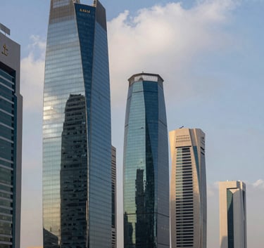 A panoramic view of a modern business district in a Middle Eastern / Gulf city during the daytime. The architecture is sleek and contemporary with glass buildings reflecting the sky blue and soft cloud white sky.