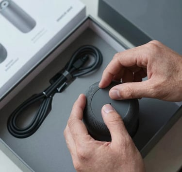 Top-down view of hands carefully opening the box of a new smart speaker, neatly organized cables and manuals visible, slate grey and light grey color palette, professional photography in a North American / US setting.