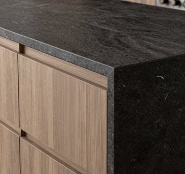 Close-up of a high-end custom kitchen island in a Southern European / Spanish (Madrid) residence, showcasing expert craftsmanship in dark charcoal stone and warm taupe wood.