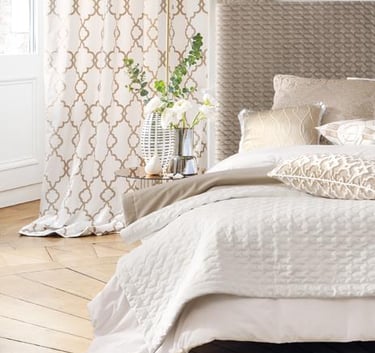 Elegant bedroom with neutral bedding, textured headboard, and pattern curtains on parquet flooring.