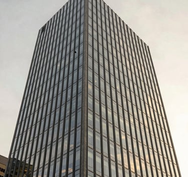 A wide-angle exterior shot of a modern glass skyscraper during the golden hour. The soft off-white sky reflects on the slate grey window panes. Professional and clean aesthetic.