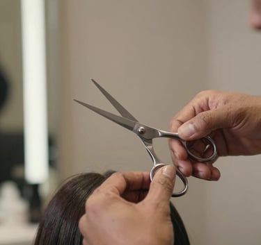 Close-up of a professional stylist working on a client's hair in a brightly lit South Asian salon, using professional silver scissors, soft focus on the modern taupe background.