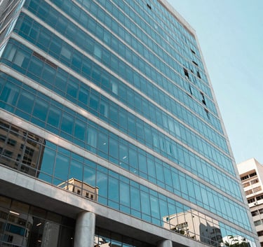 A wide-angle professional photograph of a modern Brazilian corporate building with large glass windows reflecting a cyan blue sky. The scene is bright and airy with clean lines, representing transparency and efficiency. South American urban setting.