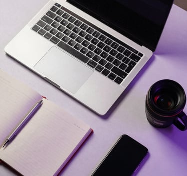 A top-down view of a modern desk with a laptop, a notebook, and a smartphone, neatly organized on a light purple surface in a professional Pakistani creative studio.