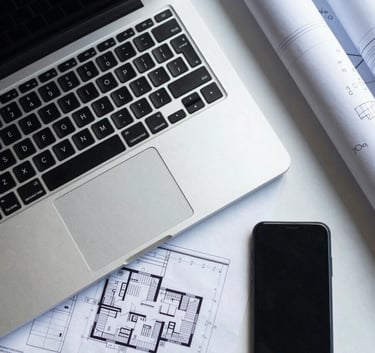 A top-down view of a professional workspace with a laptop, blueprints, and a smartphone, lit by cool morning light in Crisp Mist White and Soft Slate Blue tones.