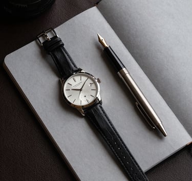 An editorial flat lay on a dark leather desk featuring a luxury watch, a high-quality steel fountain pen, and a minimal grey notebook. Soft lighting creates elegant shadows and highlights the precision of the objects.