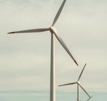 A cinematic shot of modern wind turbines standing tall against a pale off-white and soft sage green sky. The landscape is clean and well-maintained, representing sustainable and efficient energy production.