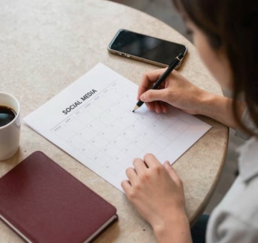 A top-down professional photograph of a marketing specialist in a cozy North American / US cafe, organizing a social media content calendar. On the Crisp Parchment table, there is a Deep Ripe Crimson notebook, a smartphone, and a cup of coffee. Warm, soft natural lighting.