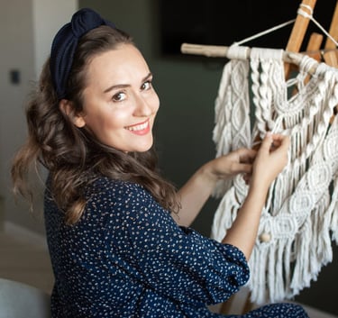 Una mujer sonriente creando un tapiz de macramé hecho a mano sobre un caballete de madera en casa.