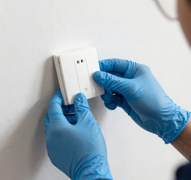 A close-up photograph of a professional technician's hands wearing protective gloves while installing a modern smart light switch. The setting is a clean Southeast Asian / Malaysian home interior. Lighting is bright and precise, featuring Soft Arctic White and Steel Blue tones.