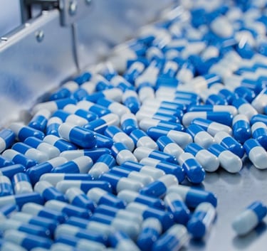 Blue and white pharmaceutical capsules moving along a stainless steel conveyor belt in a manufacturing plant.