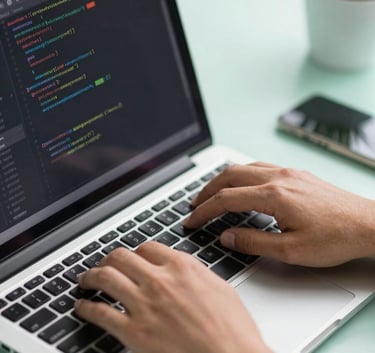 A close-up shot of a developer's hands typing on a laptop with lines of clean code on the screen. The workspace is bright and features soft mint green elements, reflecting a professional Southeast Asian / Indonesian tech environment.