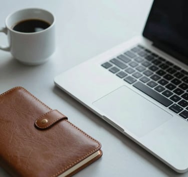 A professional desk setup featuring a sleek silver laptop, a leather notebook, and a coffee cup on a pale misty white surface, with soft slate blue lighting.