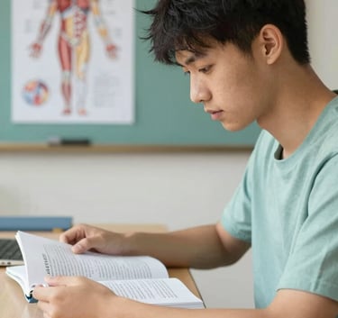 A determined student sitting at a clean desk, studying medical textbooks with a human anatomy diagram visible in the background. Soft natural light focusing on the student. Palette accents of #4F7F8F and #E0D8C0.