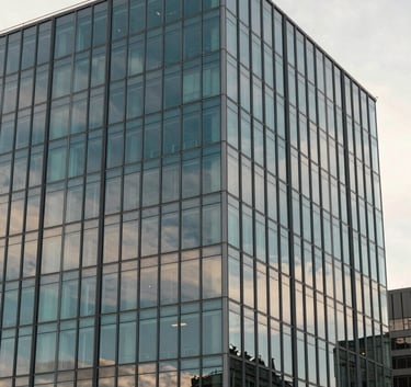 A wide shot of a modern architectural glass building reflecting a soft off-white morning sky. The image has a professional and corporate feel with muted teal and steel blue accents in the glass panes.