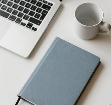 A top-down view of a clean desk in a North American / International office. A silver laptop, a steel blue grey notebook, and a soft off-white ceramic cup. Modern, minimalist, and professional.