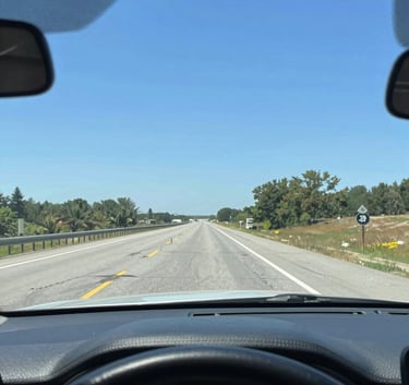 A bright, high-clarity photograph looking through a perfectly clean car windshield from the interior. The view shows a scenic North American highway stretching forward under a bright blue sky. The dashboard is clean and modern, conveying a sense of safety and a clear path ahead.